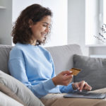 Happy hispanic young woman consumer holding credit card and laptop buying online at home. Female shopper customer shopping on ecommerce website market place making digital payment using bonus money.